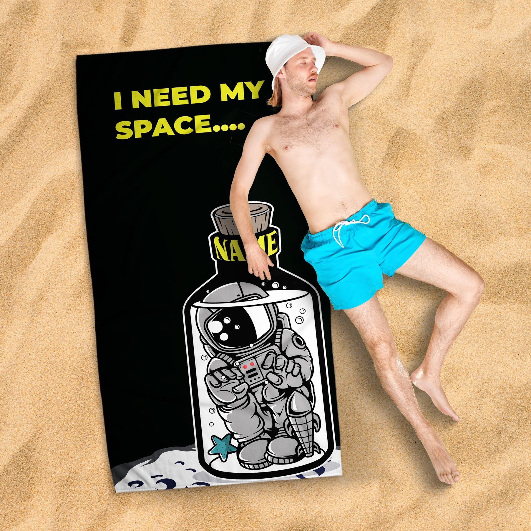 A grown man with a white hat taking a nap lying on the 'I need my space' beach towel, on a sandy surface