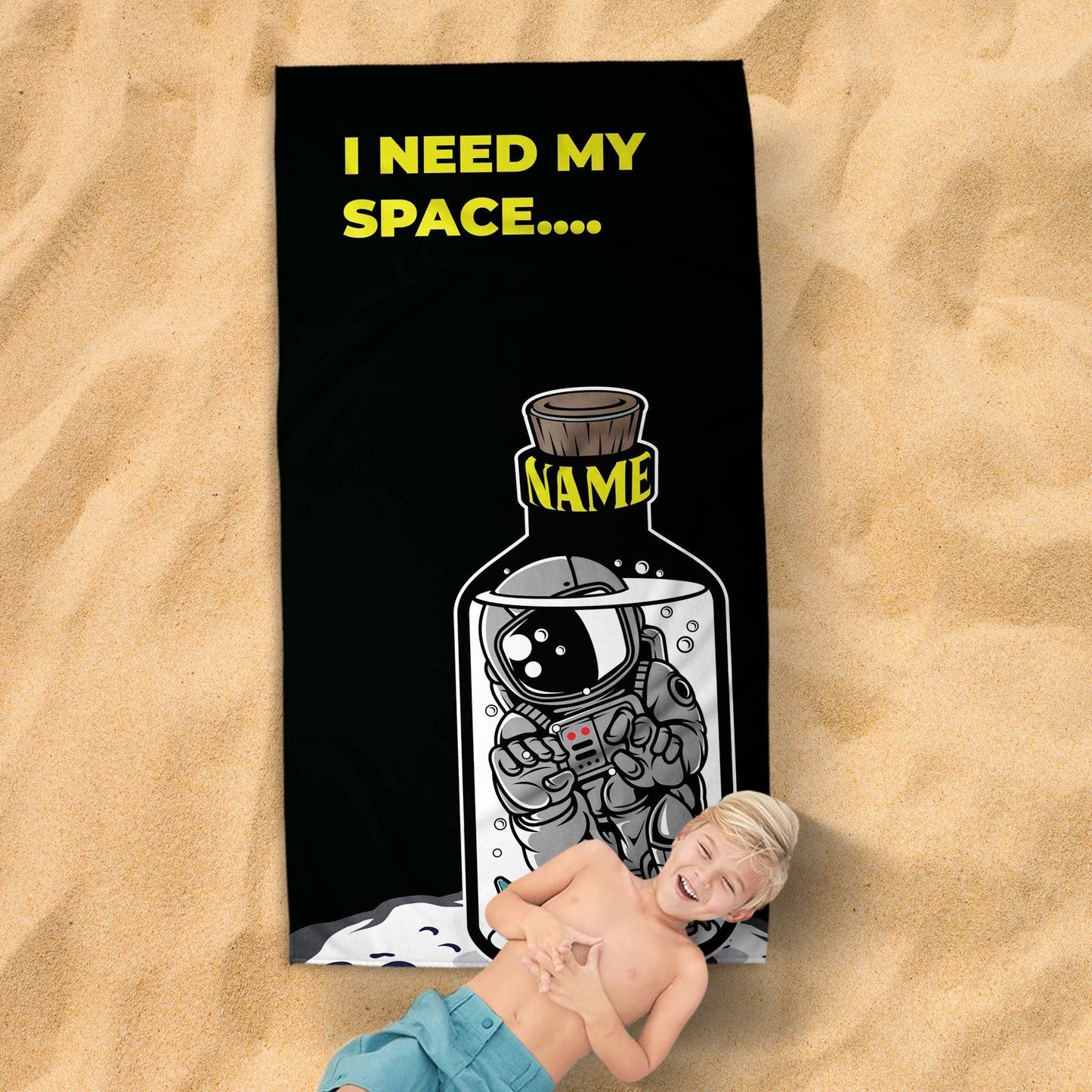 A kid with a smiling face lying on the 'I need my space' beach towel, on a sandy surface