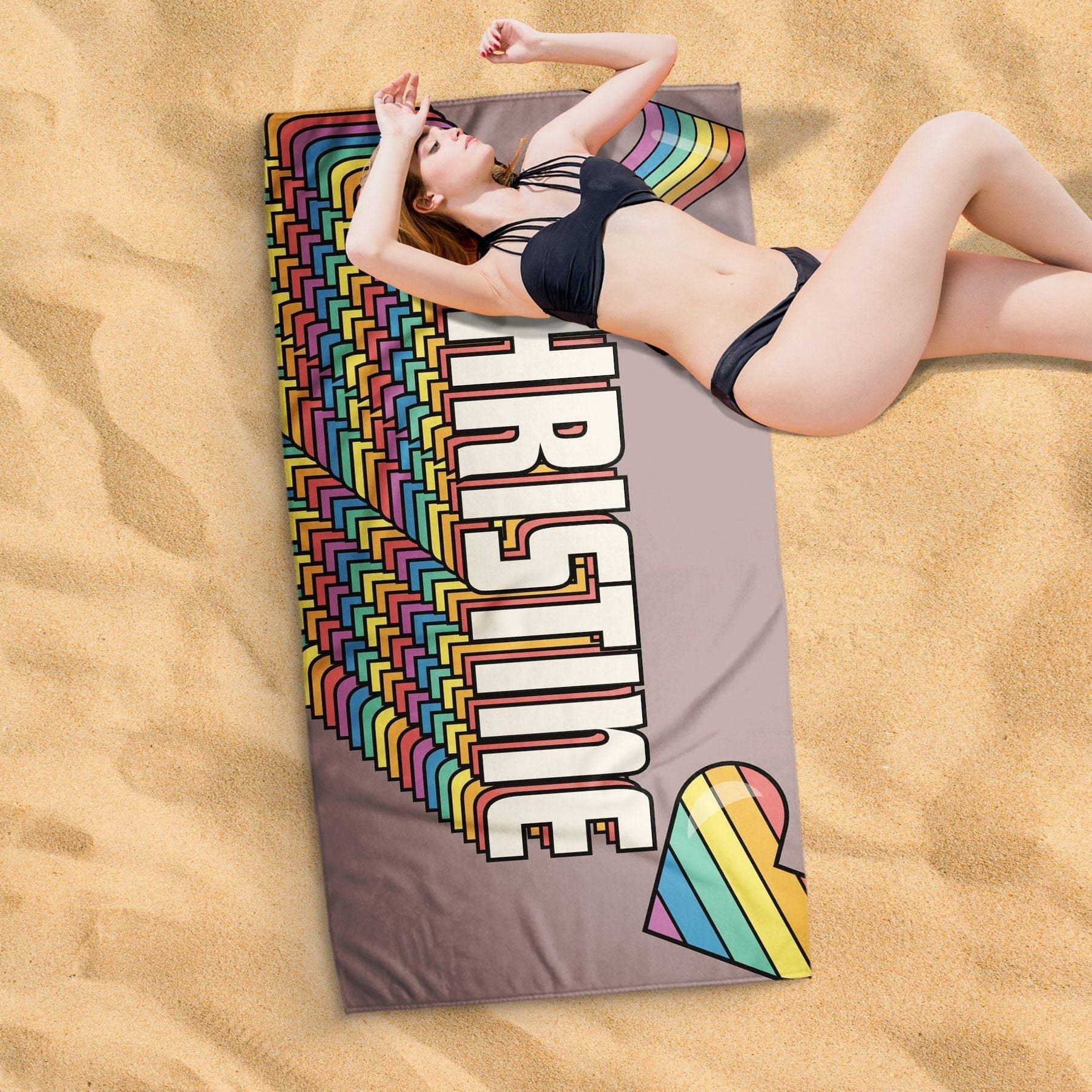 A lady lying on a retro rainbow heart beach towel on a sandy surface