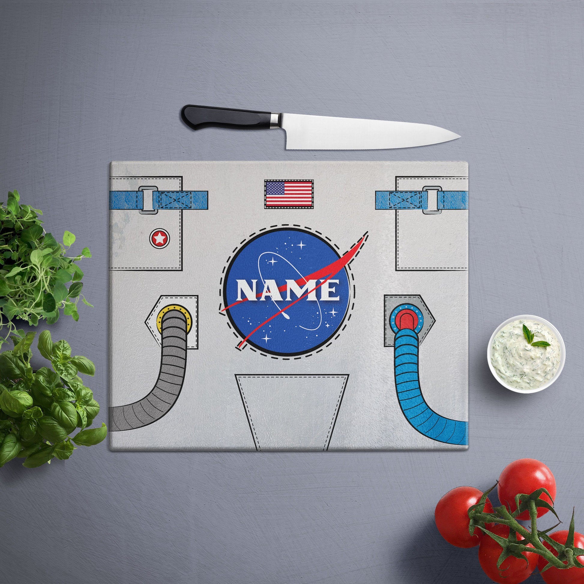 A glass cutting board with an astronaut suit design, cherry tomatoe, veggies and knife on side, on a grey surface