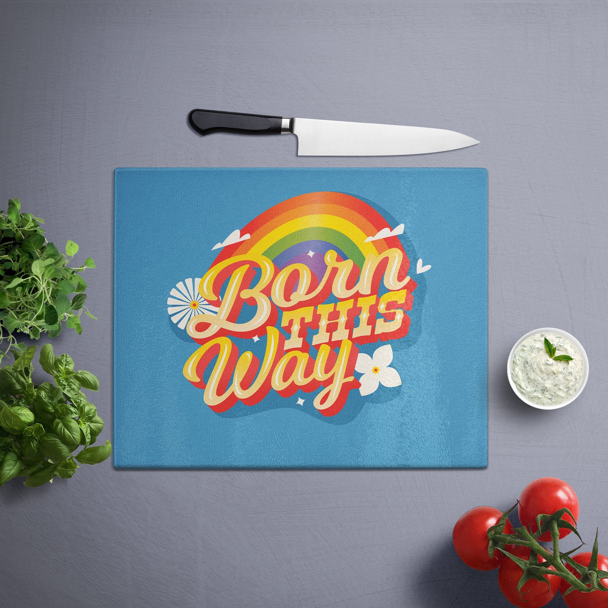 Glass cutting board with 'born this way' printed , placed on a gray surface with a knife, herbs, and cherry tomatoes nearby.