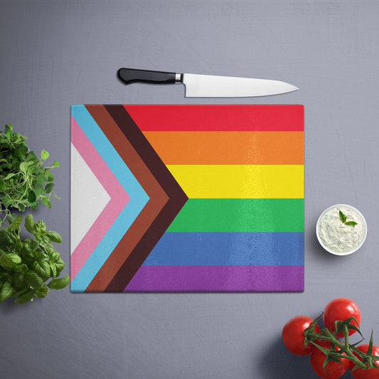 Glass cutting board with the Pride flag design, placed on a gray surface with a knife, herbs, and cherry tomatoes nearby.