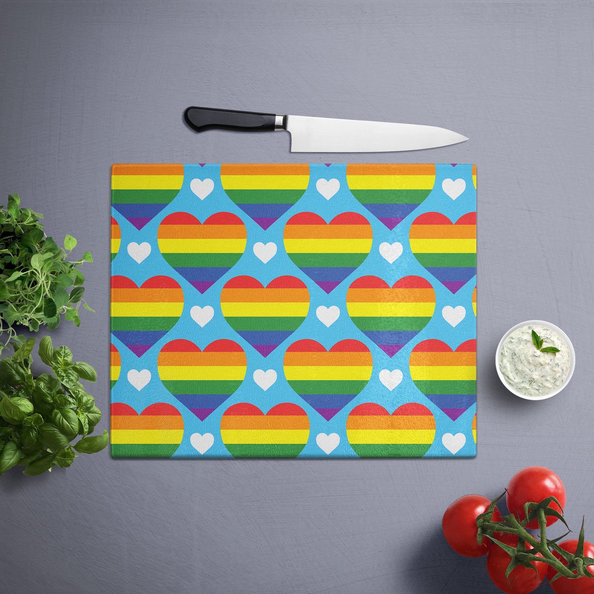 Glass cutting board with rainbow hearts pattern, placed on a gray surface with a knife, herbs, and cherry tomatoes nearby.