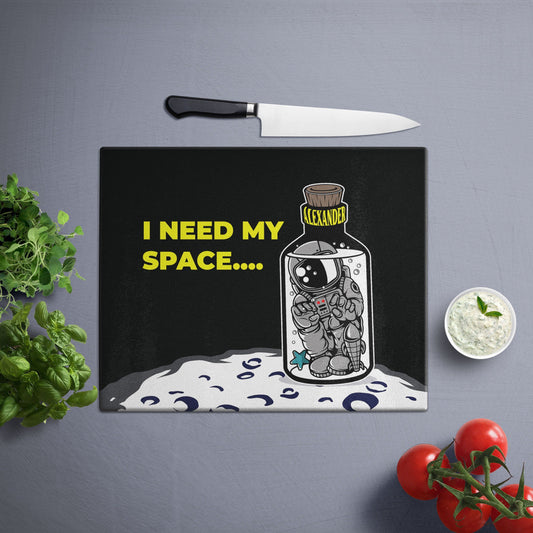 Glass cutting board with "I Need My Space" text, featuring an astronaut in a bottle labeled "Alexander," surrounded by kitchen items.