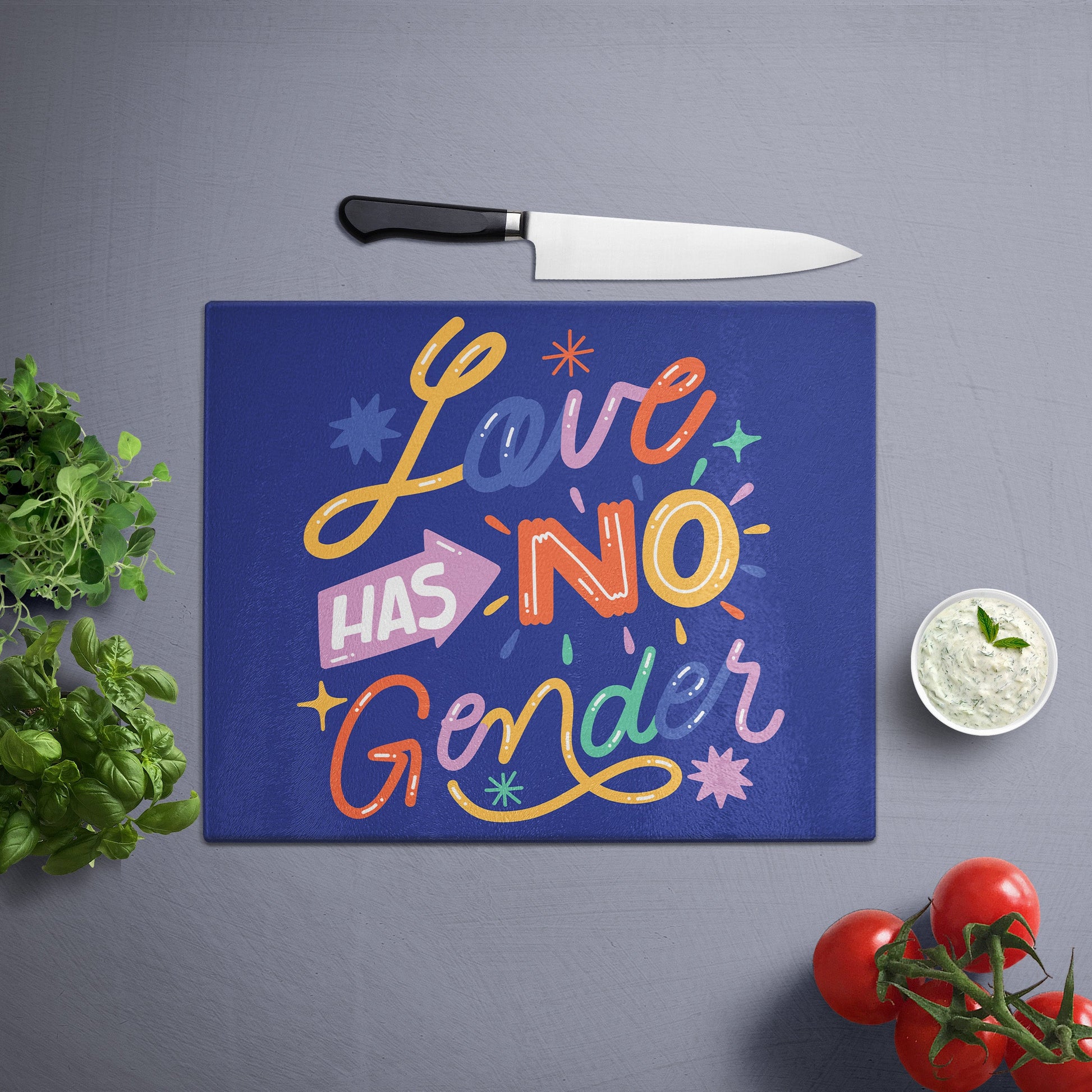 Glass cutting board with 'love has no gender' printed , placed on a gray surface with a knife, herbs, and cherry tomatoes nearby.