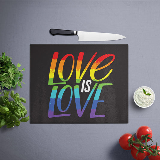 Glass cutting board with 'love is love' printed , placed on a gray surface with a knife, herbs, and cherry tomatoes nearby.