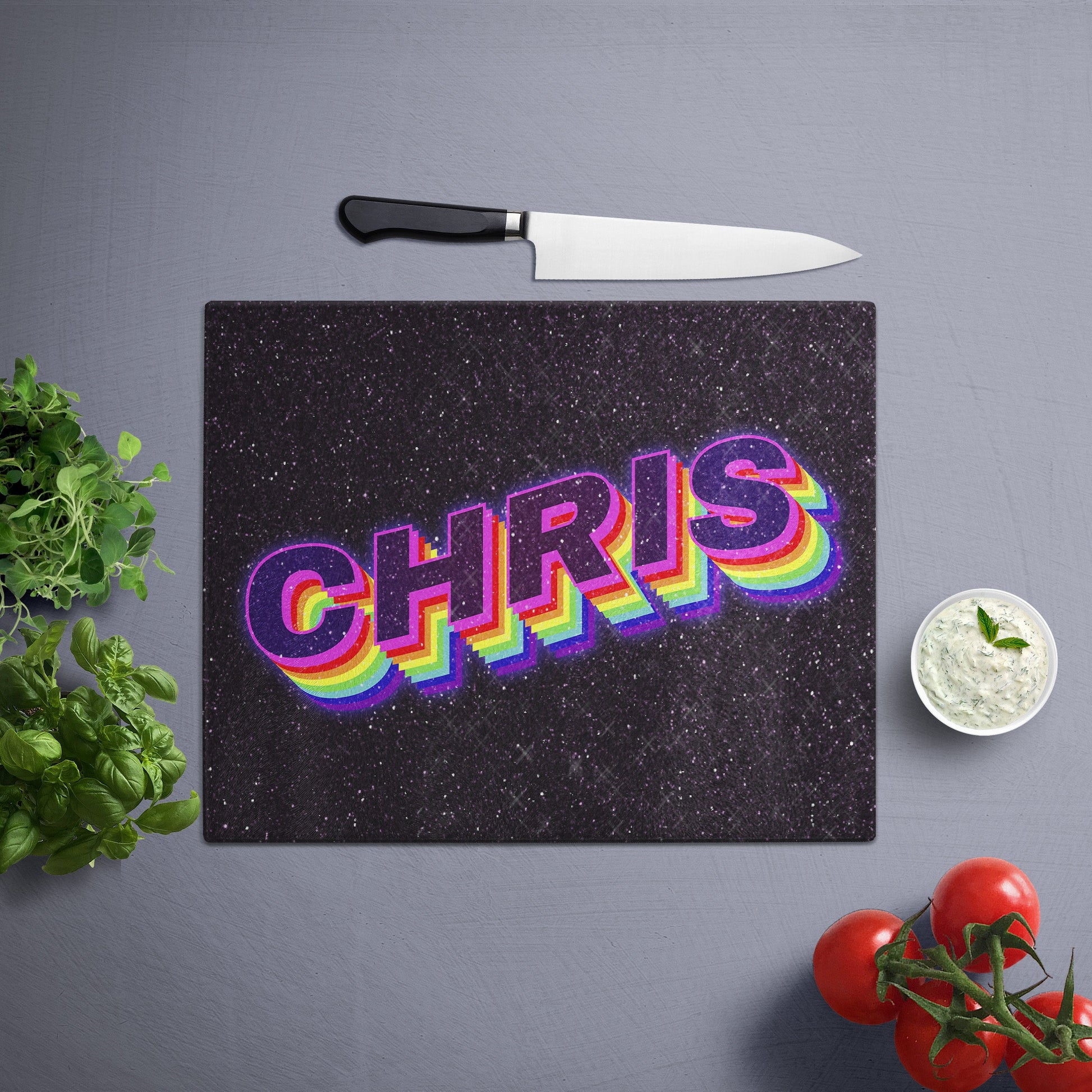 Glass cutting board with customized name in Pride flag design, placed on a gray surface with a knife, herbs, and cherry tomatoes nearby.