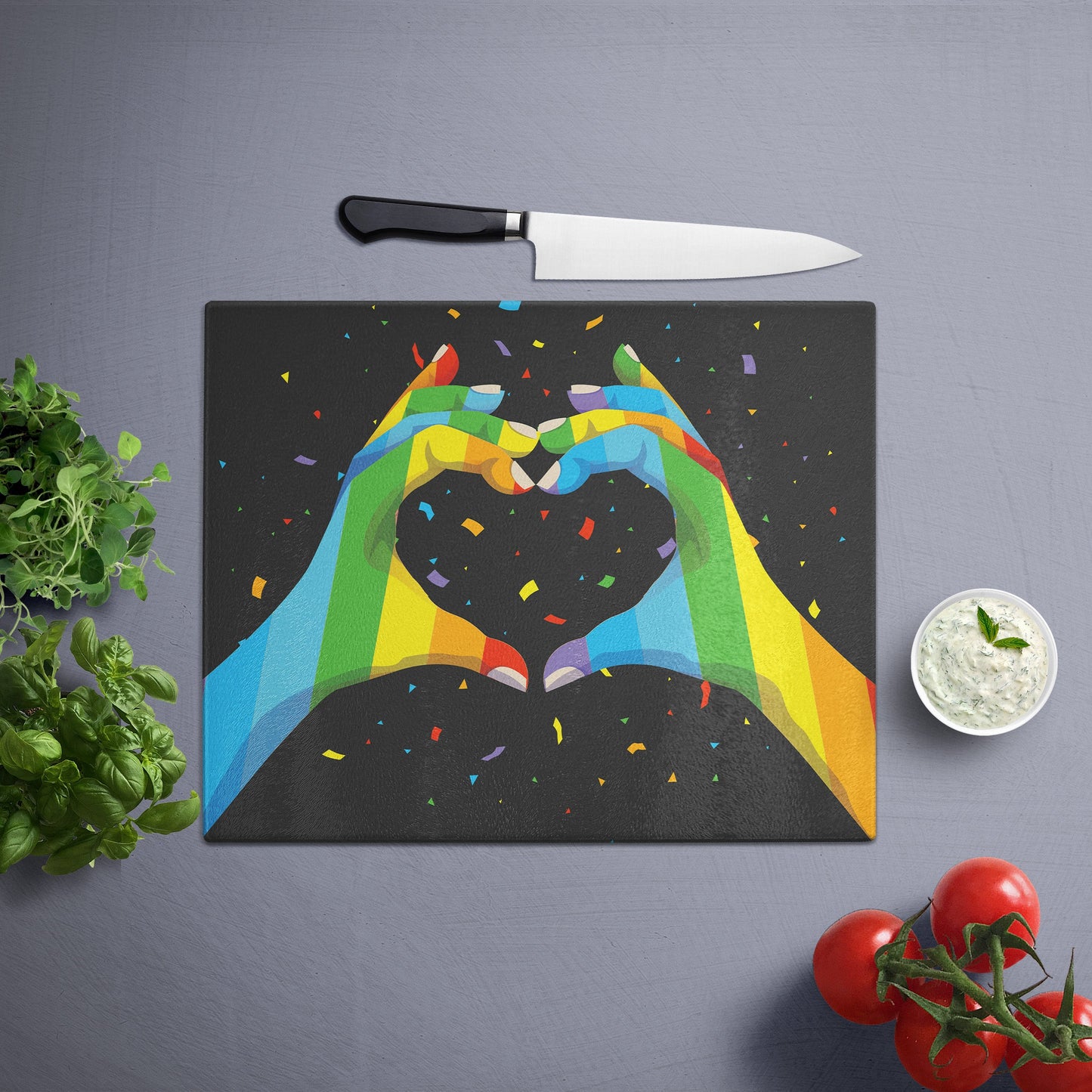 Glass cutting board with 'hands and heart' print in a pride pattern, placed on a gray surface with a knife, herbs, and cherry tomatoes nearby.