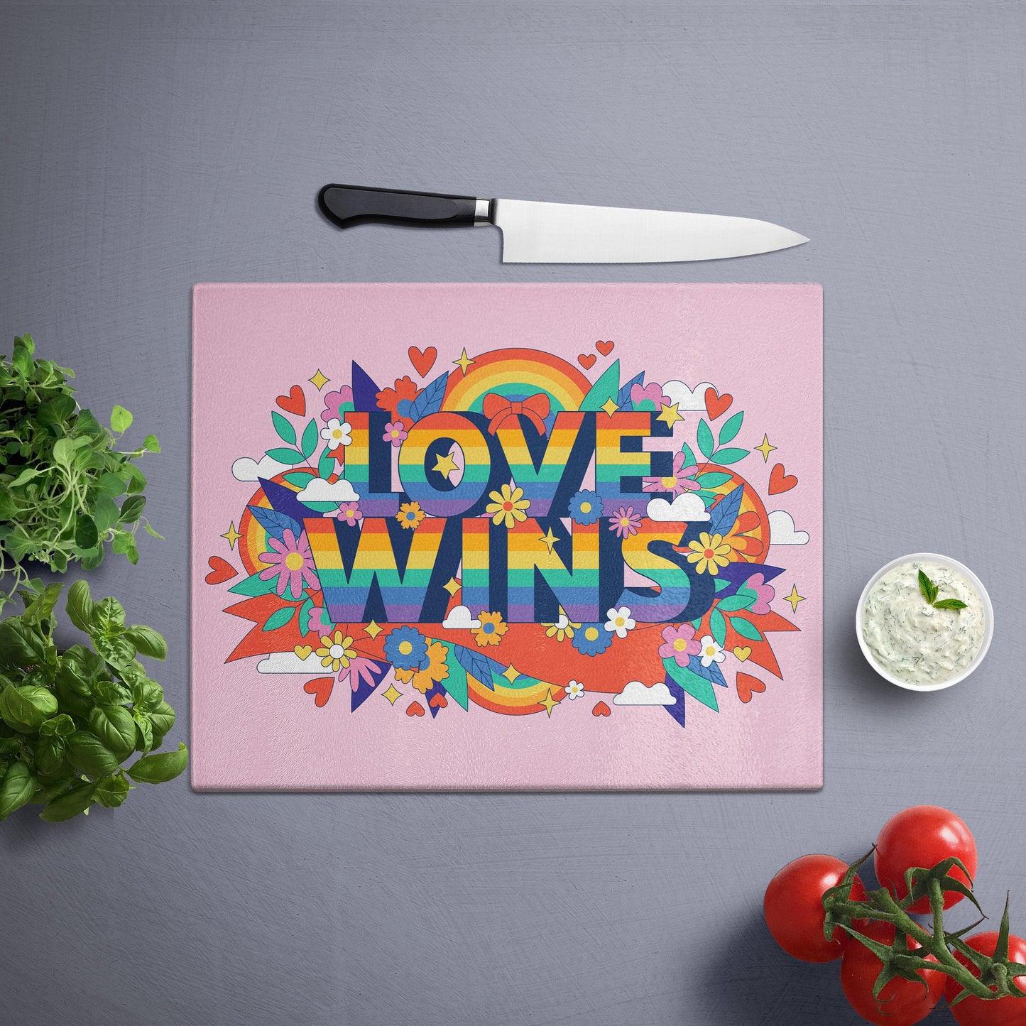 Glass cutting board with 'Love wins' print in a pride pattern, placed on a gray surface with a knife, herbs, and cherry tomatoes nearby.