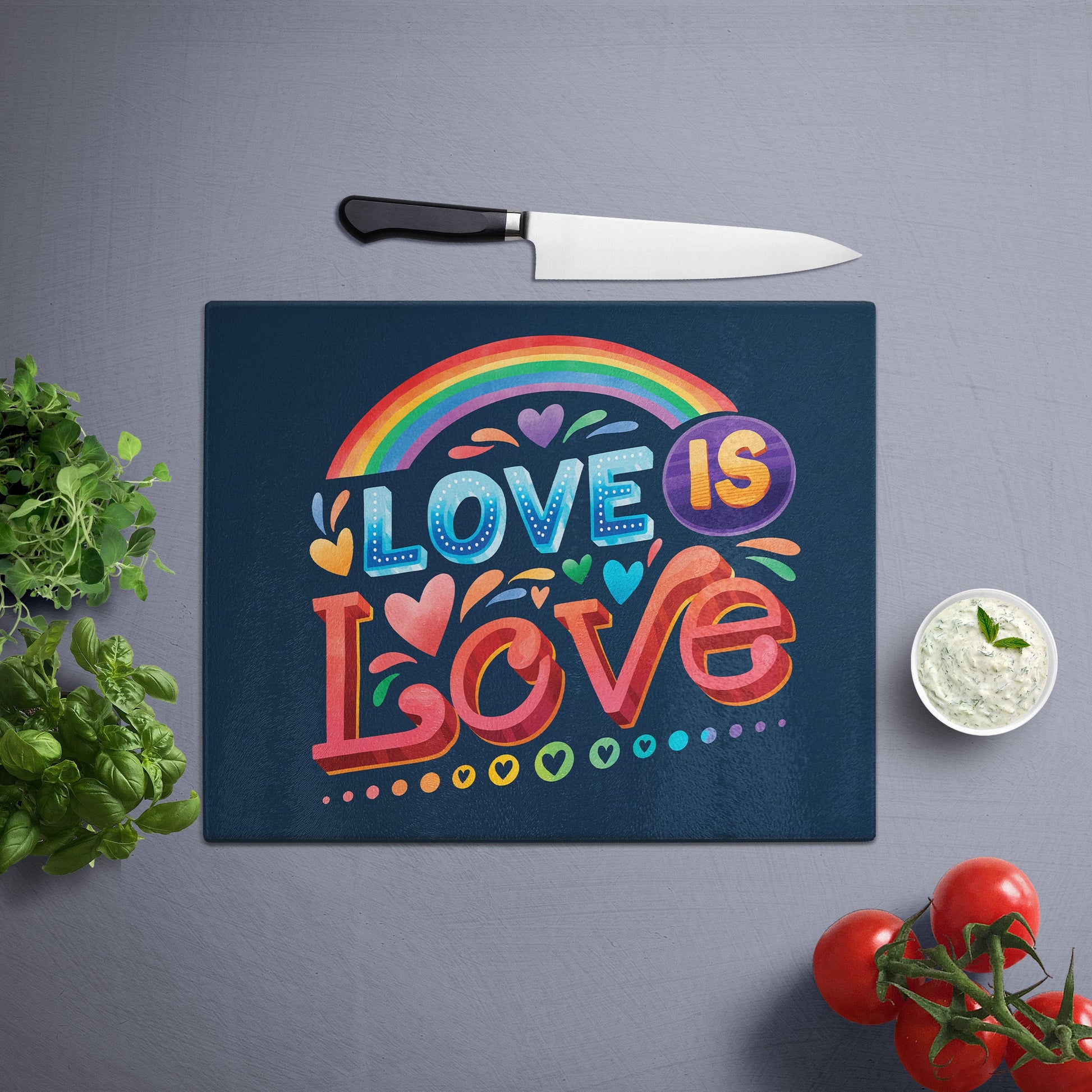 Glass cutting board with 'love is love' printed , placed on a gray surface with a knife, herbs, and cherry tomatoes nearby.