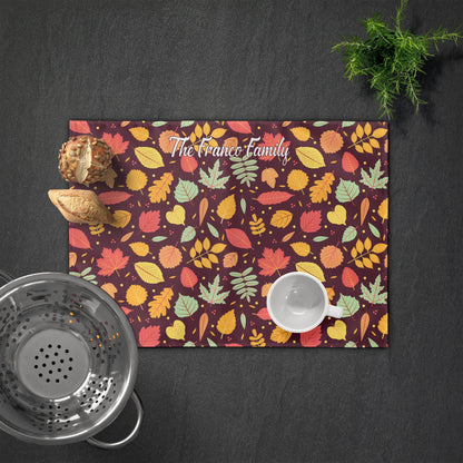 Personalized placemat with "The Franco Family" text, fall leaves pattern, and bread, colander, cup, and a small plant on a dark surface.