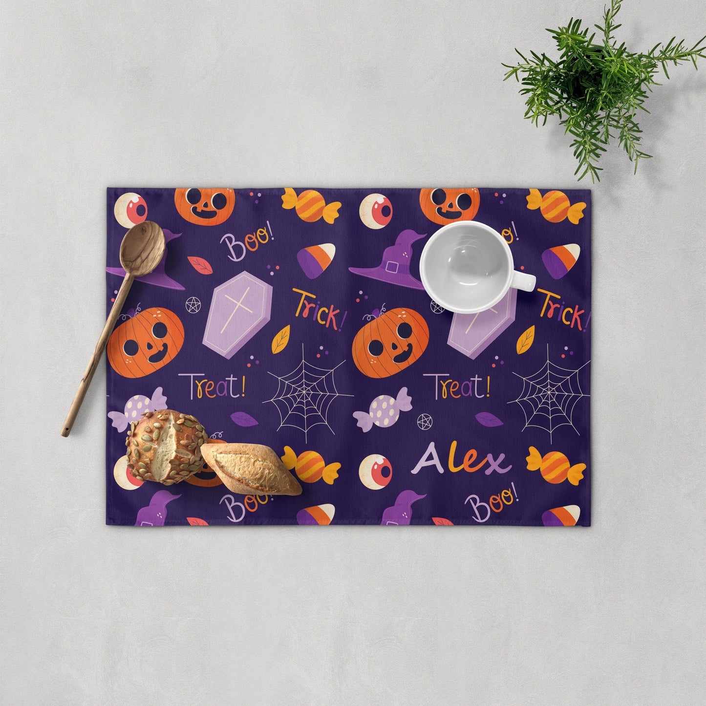 Personalized purple placemat with "Alex," featuring orange pumpkin, coffin, and candy, Bread, cup, and colander are nearby